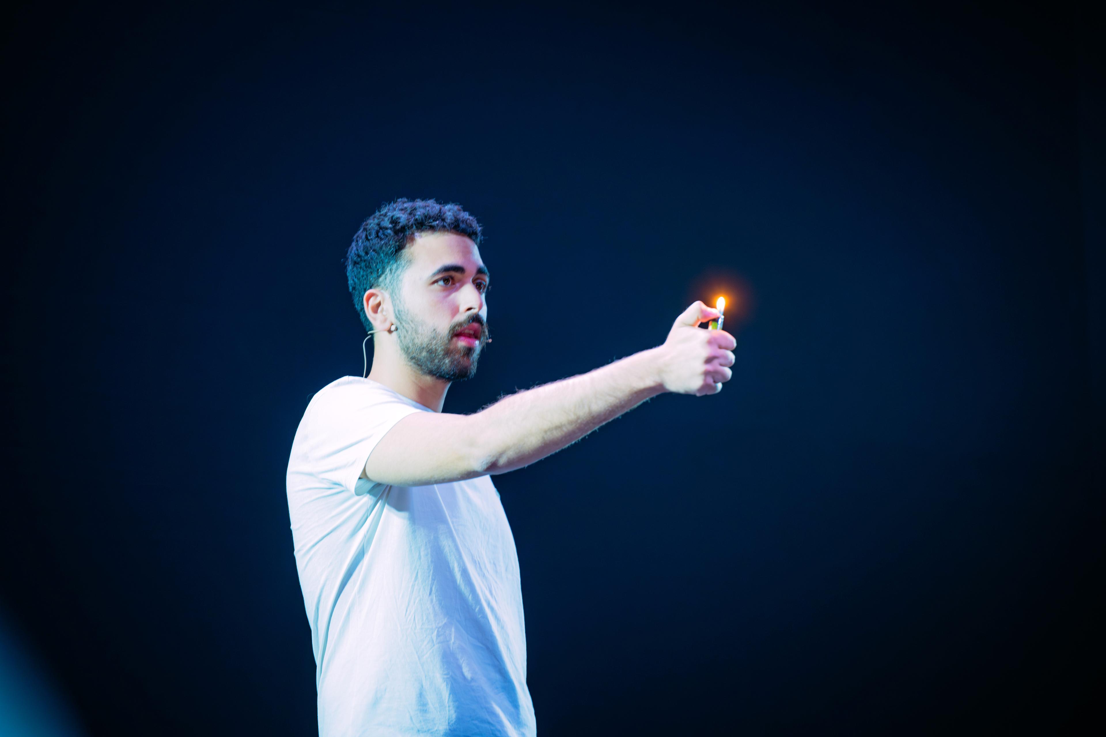 Fadi Murad on a dark stage in a white T-shirt, holding a lit lighter at arm’s length.