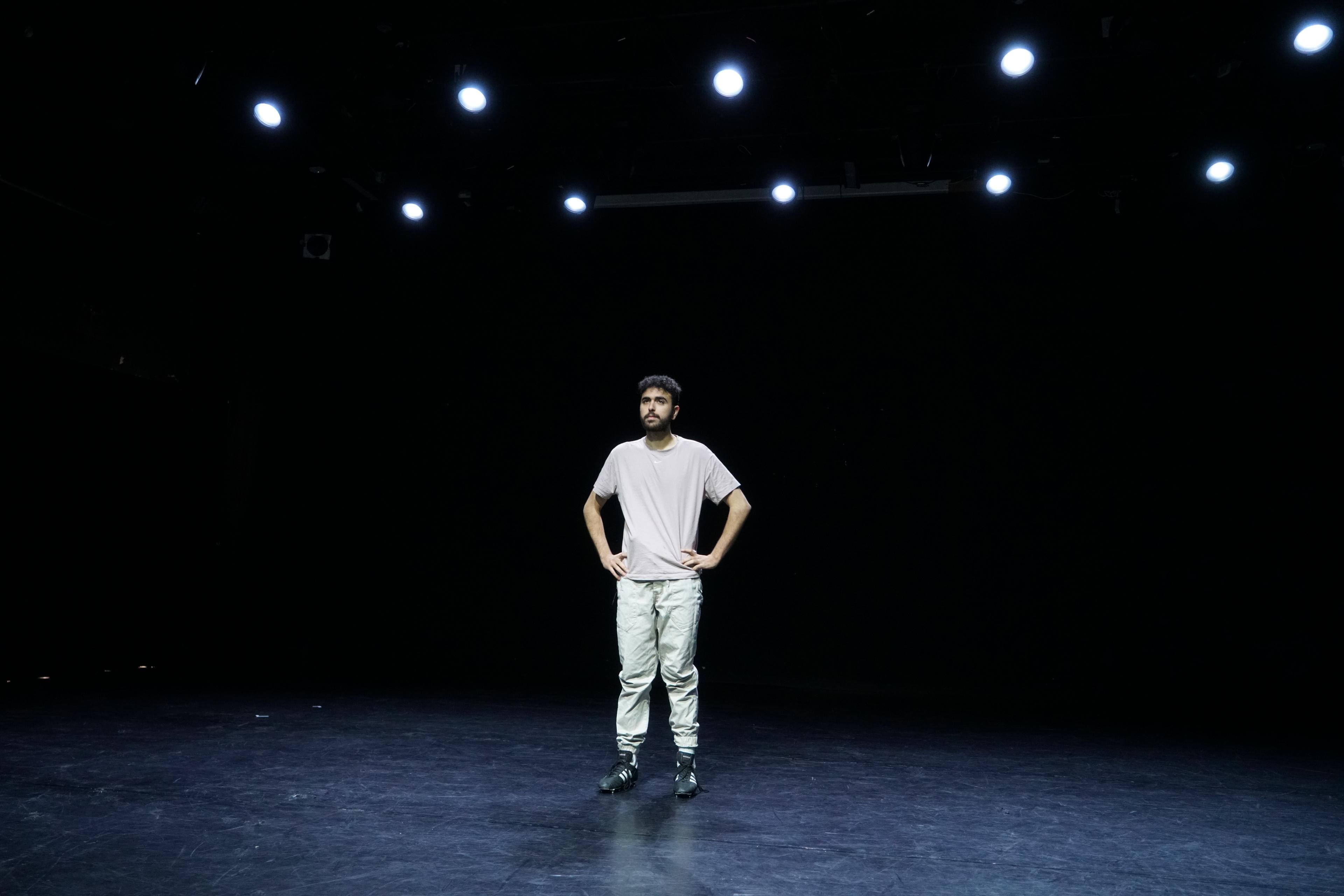 Fadi Murad stands alone, hands on hips, on a dark empty stage under a grid of bright lamps.